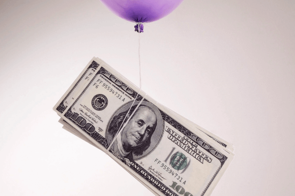 Banconote da 100$ legate da un filo di spago sono sollevate in aria da un palloncino lilla